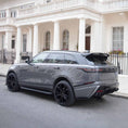 Load image into Gallery viewer, Rear quarter angle of Nardo Grey Velar Huntress parked near a white luxury hotel, showcasing bumper and diffuser styling.