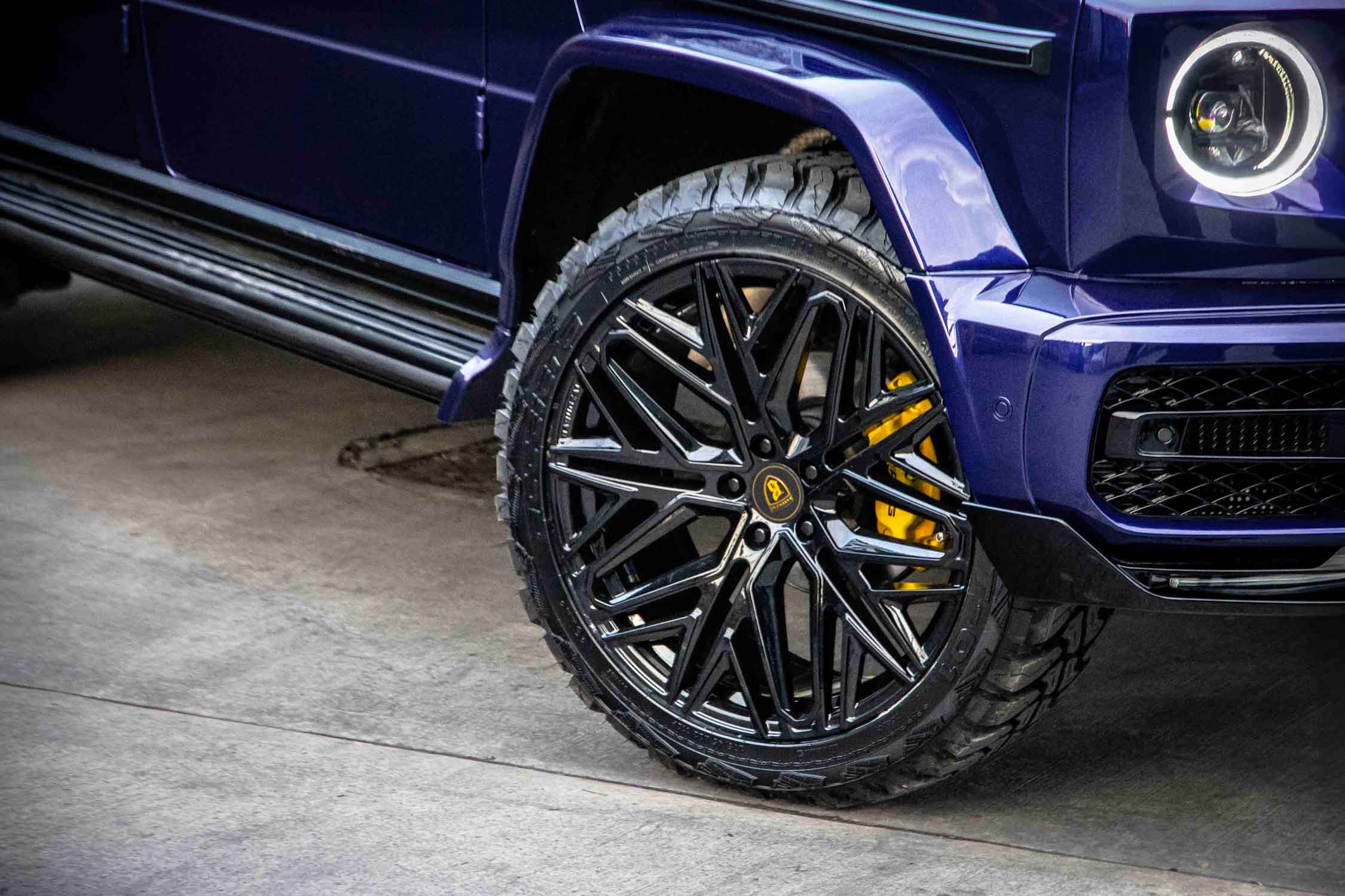Close-up of a blue Mercedes G Wagon W463A with a black Barugzai Shuracan gloss black alloy wheel with yellow calipers on a concrete surface.