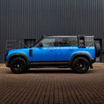 Load image into Gallery viewer, Blue SUV with black roof rack parked on a brick road in front of a dark building.
