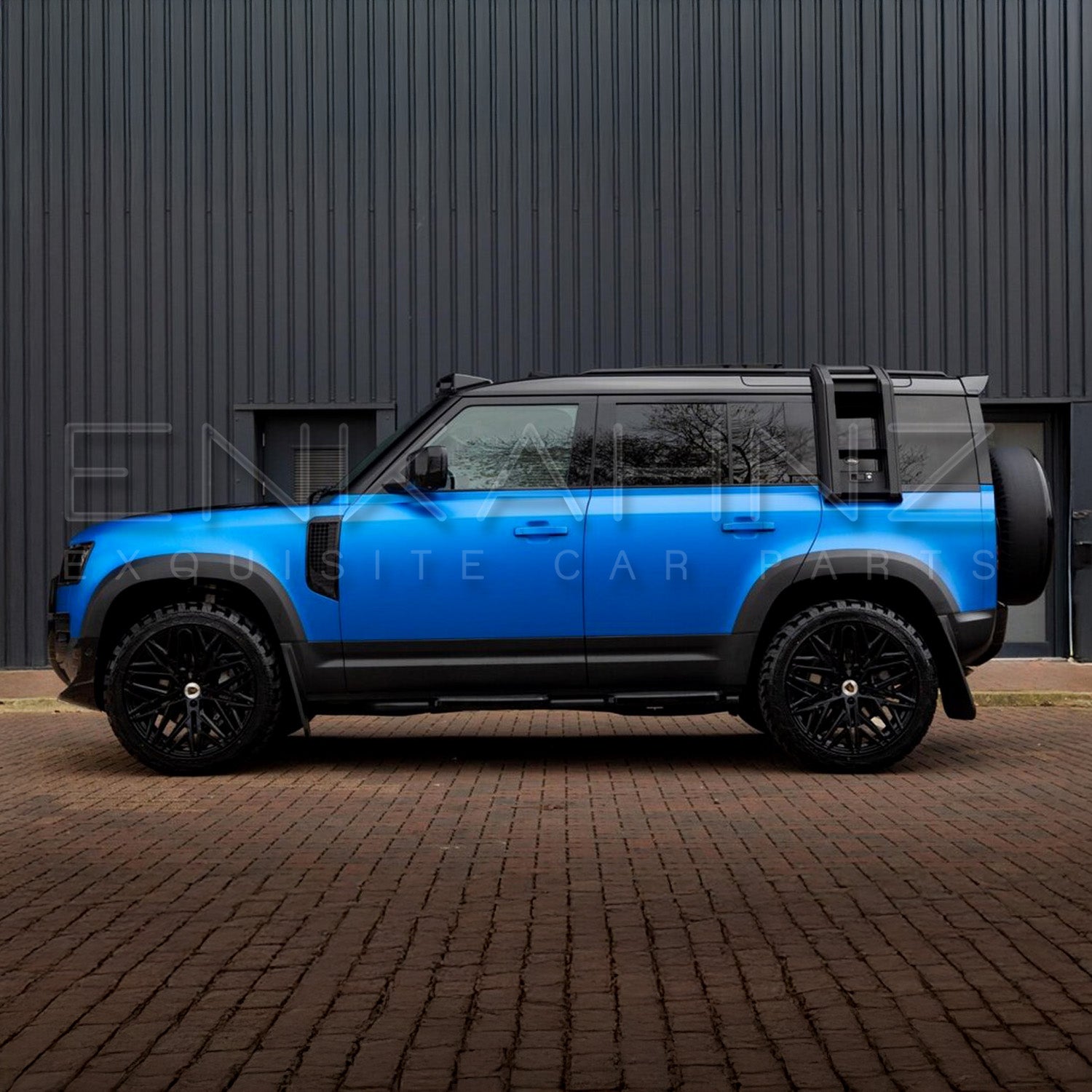 Blue SUV with black roof rack parked on a brick road in front of a dark building.