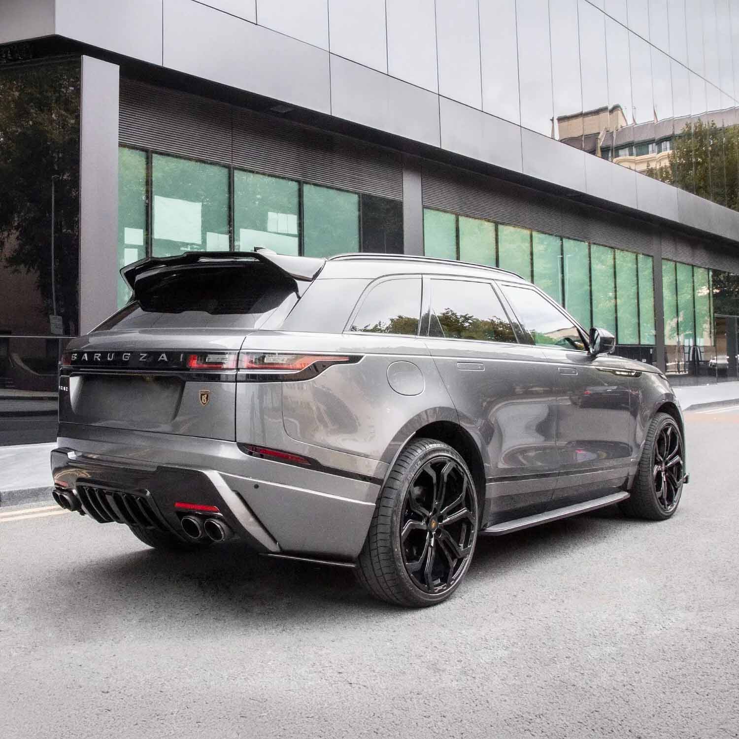 Rear slightly lower quarter angle before glass‑fronted building with green tint, showing diffuser and exhaust surrounds.