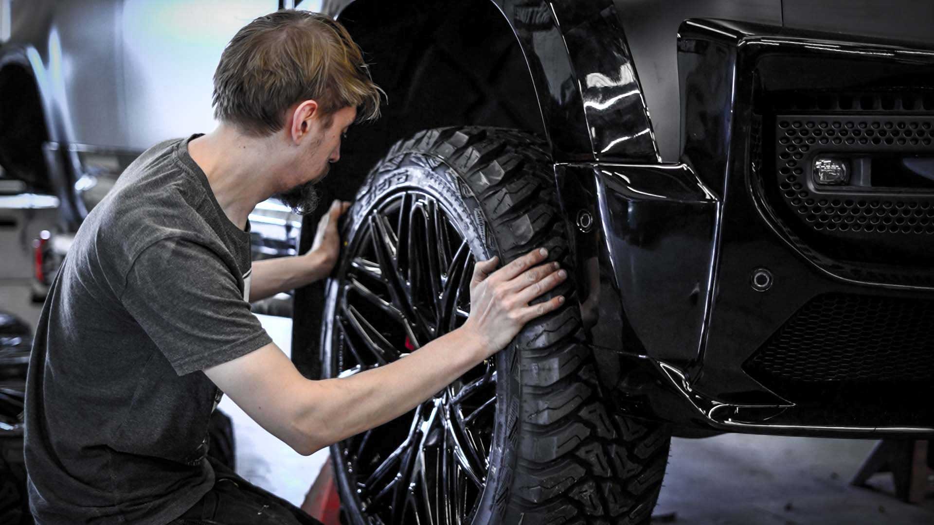 Person fitting Barugzai's Shuracan 24" alloy wheel in gloss black, with all terrain tyres, on a Land Rover Defender 110 L663, Barugzai Falcon bodykit. 